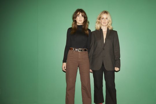 Both members dressed in neutral brown and black clothing stand in from of green wall