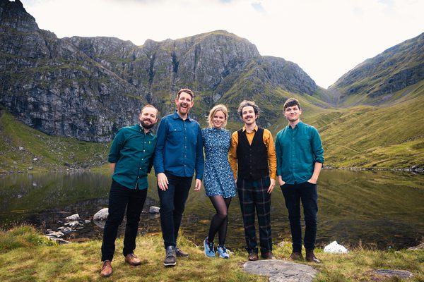 Four men and one woman, wearing different shades of green and blue, standing next to a lake in the mountains.
