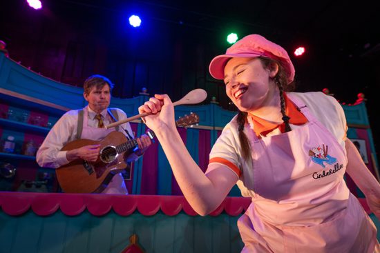 A woman in an apron and hat singing into a wooden spoon. A man is behind her playing the guitar.