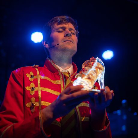 A man in a red and gold jacket with his eyes closed, holding a glass slipper.