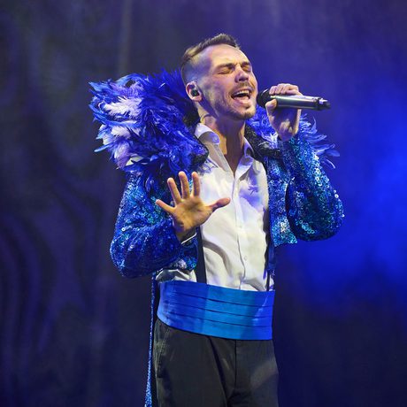 A man singing, dressed in a blue jacket with a large feathered collar.