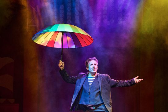 Man standing under colourful lights holding a rainbow umbrella above his head.
