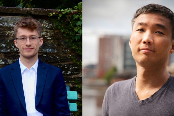 Two men. On the left is a man wearing glasses sat on a blue bench. On the right is a man closer to the camera in a grey t-shirt.