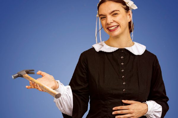 A lady in a black dress with a white collar holding a hammer and smiling.