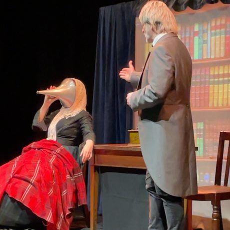 A blonde man talking to a woman with a cone over her face and a red blanket over her knee.