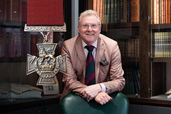 Mark Smith sitting in a library next to a large image of the Victoria Cross.