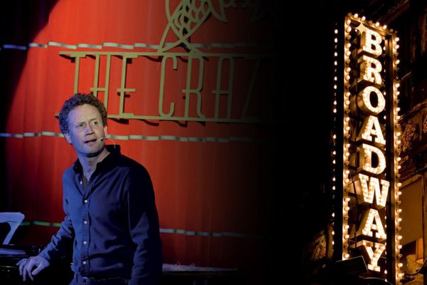 Man in a black shirt standing in front of a red curtain with a lit up Broadway sign next to him.