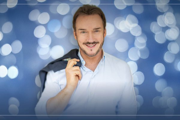 Russell Watson standing in front of a blue sparkly background, holding his jacket over his shoulder.