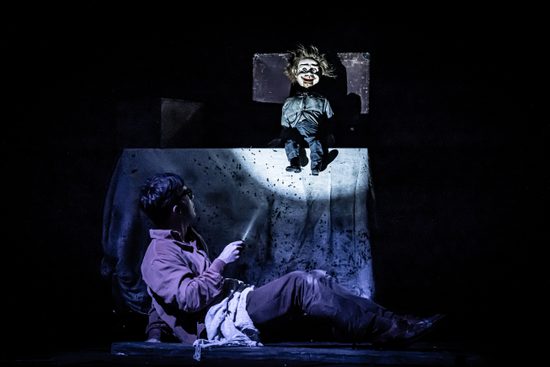 A man sitting on the floor, shining a torch on a creepy doll.