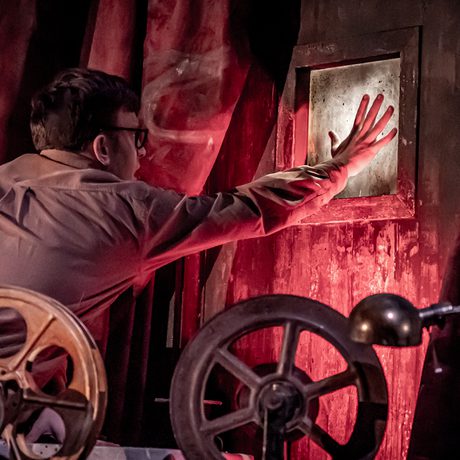 A man in brown clothes surrounded by red lighting reaching out towards a window.