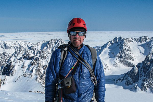 Simon Yates wearing a blue coat and red helmet, standing on top of a snowy mountain.