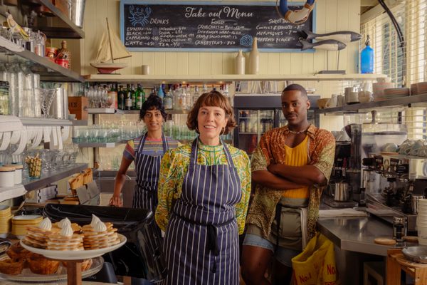 Olivia Coleman standing in front of two other people in a kitchen, wearing blue and white striped aprons.