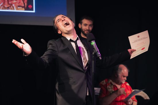 A man in a suit standing at a microphone, laughing with a piece of paper in his hand.