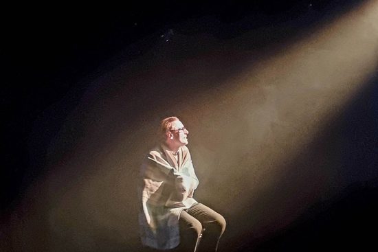 A man sat on a chair, wrapped in a blanket, under a spotlight.