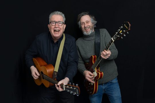 Tom Robinson and Adam Phillips standing with guitars and smiling.