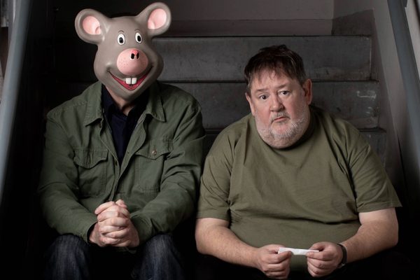 man in a mouse mask and Johnny Vegas sit at the bottom of a set of concrete stairs. Vegas is rolling a cigarette