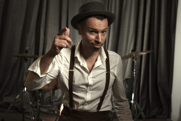 Si in a white shirt and black hat and suspenders tapping his finger to the music.