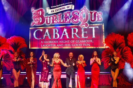 Dancers wearing red feathers and singers in a line on a stage, smiling.