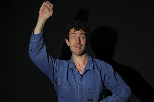 A man in a blue jumpsuit with one arm above his head.
