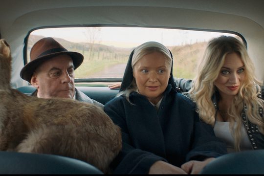 A man in a brown hat, a nun, and a woman in a leather jacket sit in the back of a car.