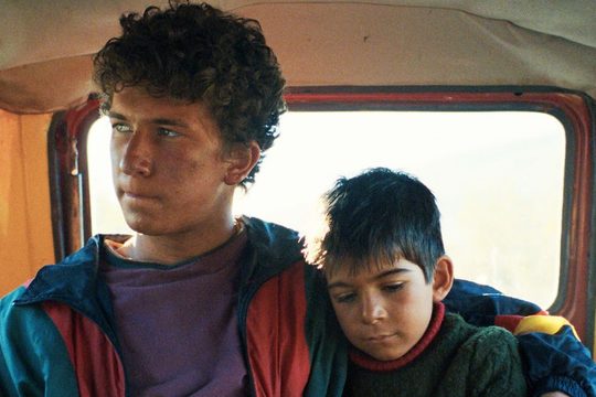 Two boys, one a teenager and one a younger child, sit in the back of a vehicle. The older boy has his arm around the younger.