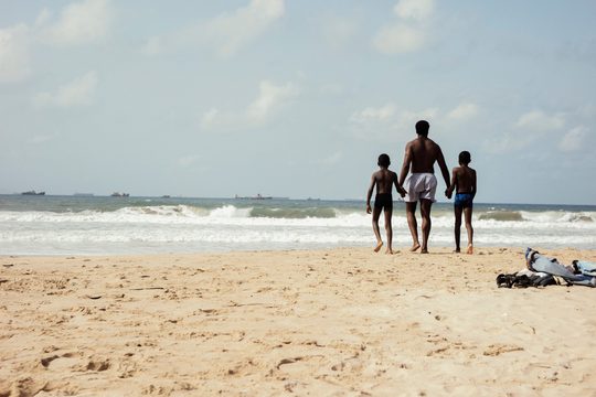 A man holding hands with two younger boys. They are all wearing swimming trunks and are on the beach, looking out to sea.