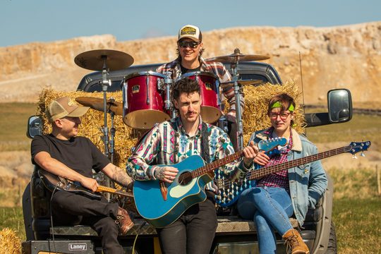 Four band members playing various instruments including guitar, bass guitar, and drums. They are sat in the back of a van that is stacked with hay bails.
