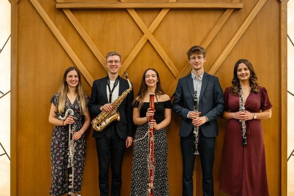 Five musicians in a line all holding instruments. From left to right, there is a bass clarinet, a saxophone, a bassoon, a clarinet, and an oboe.