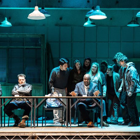 A stage lit in team with a long table across the front. A man speaks into a microphone and various others lean over him.