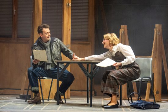 A blonde woman in a tartan jumpsuit and white shirt holds hands across a table with a brunette man waring a leather jacket and a grey scarf.