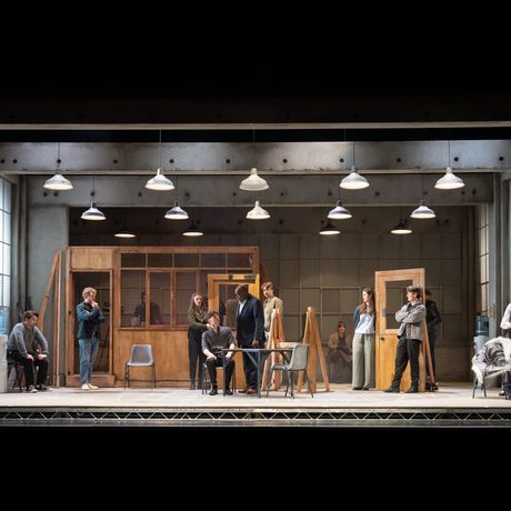 A concrete stage with multiple pieces of simple wooden set. There are various people sat around the stage, looking at a man sat in a plastic chair.