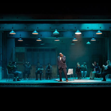 A stage lit in a teal colour. There are multiple people scattered around the stage sat on plastic chairs, and multiple overhead lights.