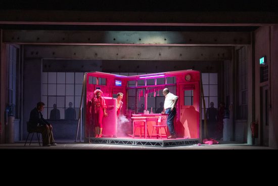 A bare stage with a bright pink set in the middle, exhibiting a kitchen.