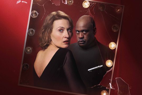 A mirror with lights around it set on a red background. The mirror shows a man and a woman, both dressed in black and looking scared.