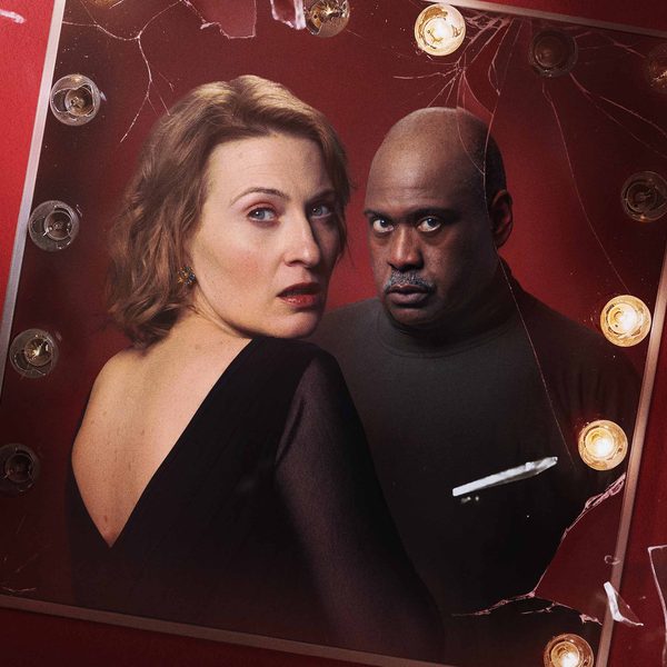 A mirror with lights around it set on a red background. The mirror shows a man and a woman, both dressed in black and looking scared.