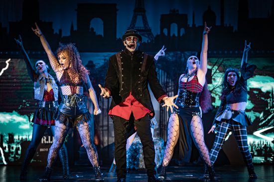 A man in a red shirt, black jacket and black hat singing with four women singing behind him.