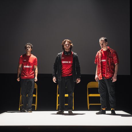 Three men in red football shirts standing in a spotlight in front of metal yellow folding chairs.