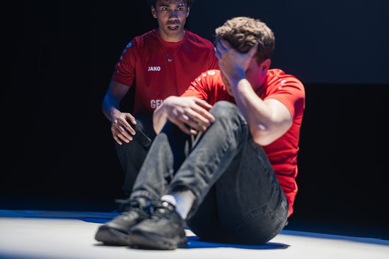 Two men in red football shirts are in a spotlight. One is curled up and looks upset, and the other is trying to comfort him.
