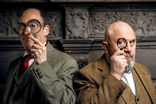 Two men in tweed jackets hold magnifying glasses to one of their eyes.