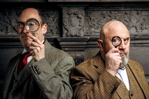 Two men in tweed jackets hold magnifying glasses to one of their eyes.