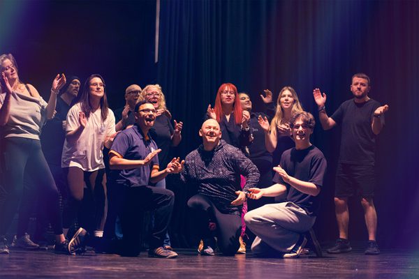 Various people wearing comfortable clothing rehearsing. They are all smiling and clapping and are stood or crouched in a group.