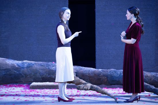 Two women stand in front of a fallen tree talking to each other. One is wearing a black top and white skirt, and is pointing to the other, in a deep red dress.
