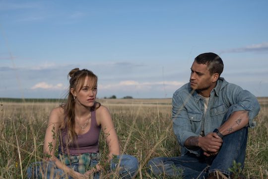 A blonde woman in a purple top and jeans and a man in a denim jacket sit cross legged in long grass.