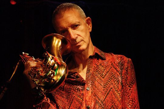 A man in a red snake-skin shirt holding a saxophone to his cheek.