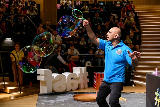 A man in a blue t-shirt creates large bubbles in front of an audience.