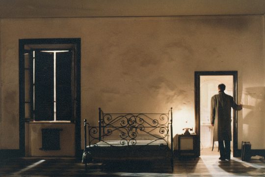 A man in a long coat stood in the doorway of an empty room with cream walls.