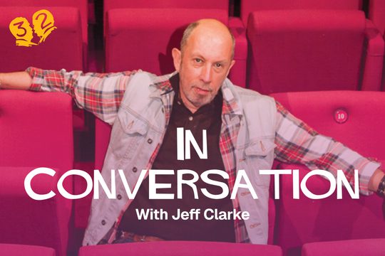 A man in a tartan shirt sitting on red theatre chairs.