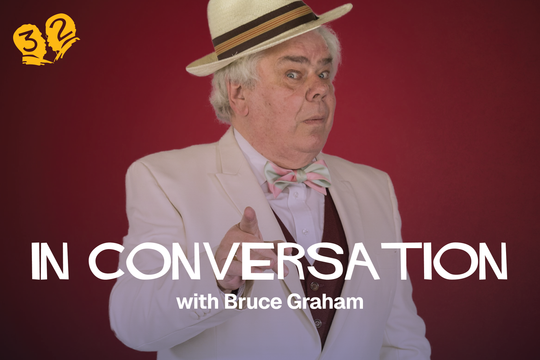 A man in a white suit, green and pink bow tie, and straw hat pointing at the camera.