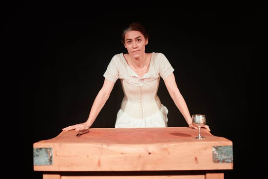 A white, brunette woman wearing a white dress stands with both hands resting on a wooden table.