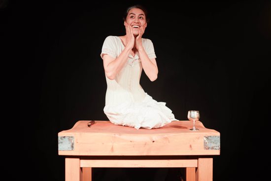 A white, brunette woman sat on a wooden table wearing a white dress. Both her hands are pressed to her cheeks and she is smiling.